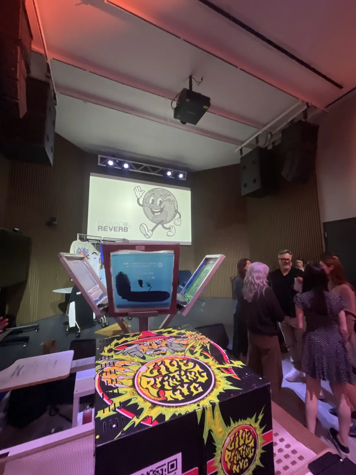 The Live Printing NYC branded press — wrapped in the wild green sun artwork — set up with screens raised at a Bravado x Reverb event, guests gathered around